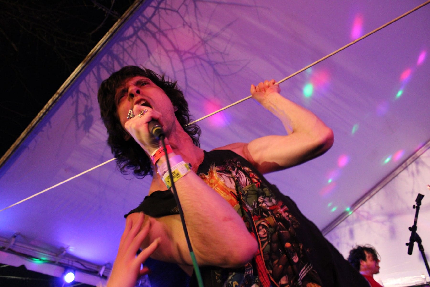 A singer leans toward the audience under purple light, an arm raised beside a diagonal microphone cable.